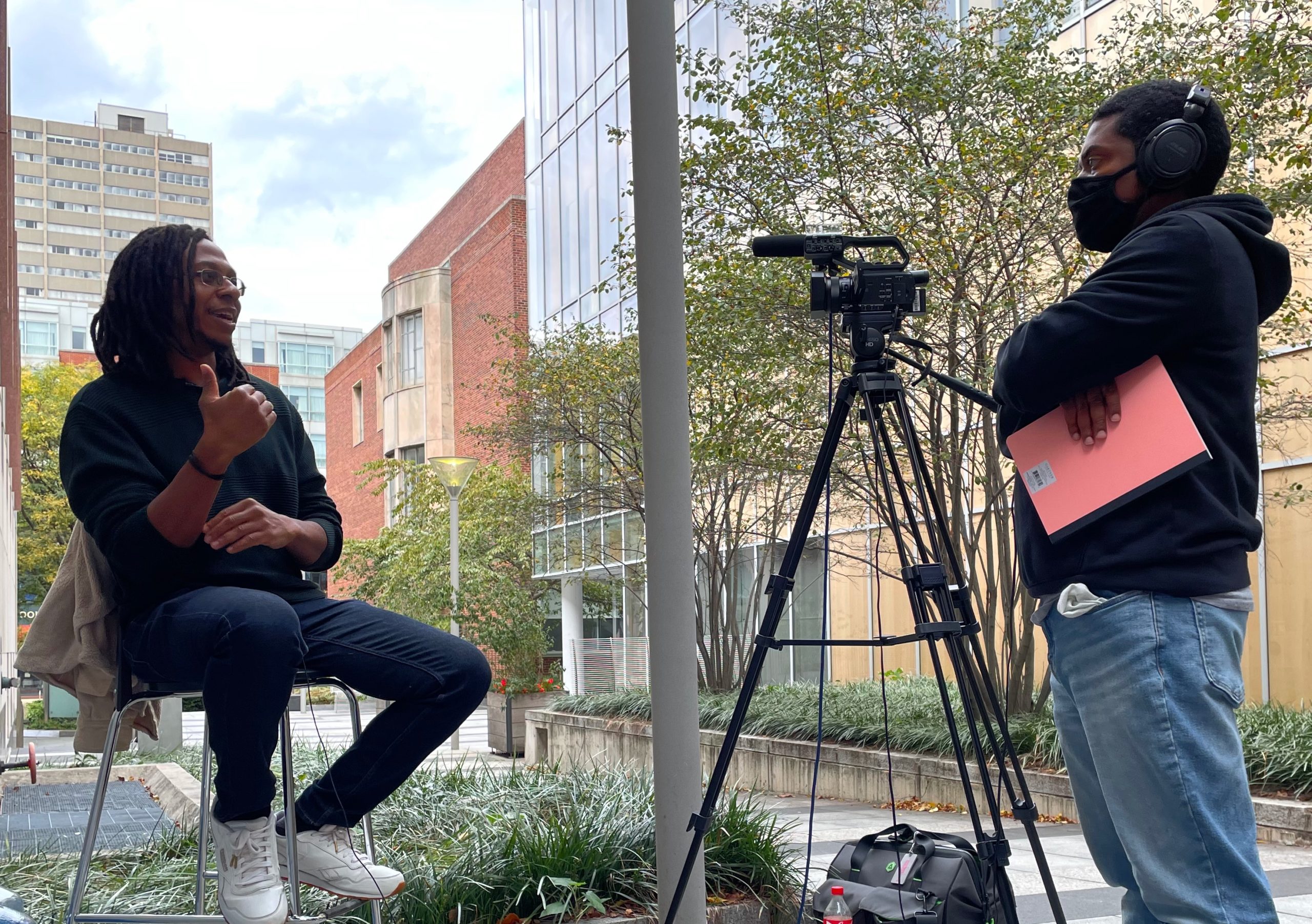 A person on the right is conducting an interview with a camera and mic on a tripod, while the interviewee is seated on the left.