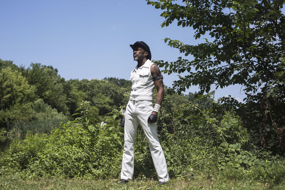 A mature black male stands proud in nature wearing a white, short sleeved leather shirt and white leather pants. He is accessorized with a black leather western hat and a white studded thick wristband.