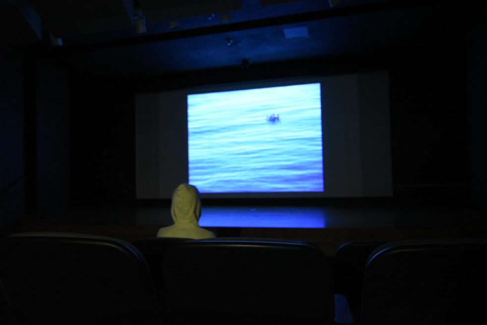 a hooded figure sitting in a dark room, watching a large screen on which appears a body of water and in the distance a boat.