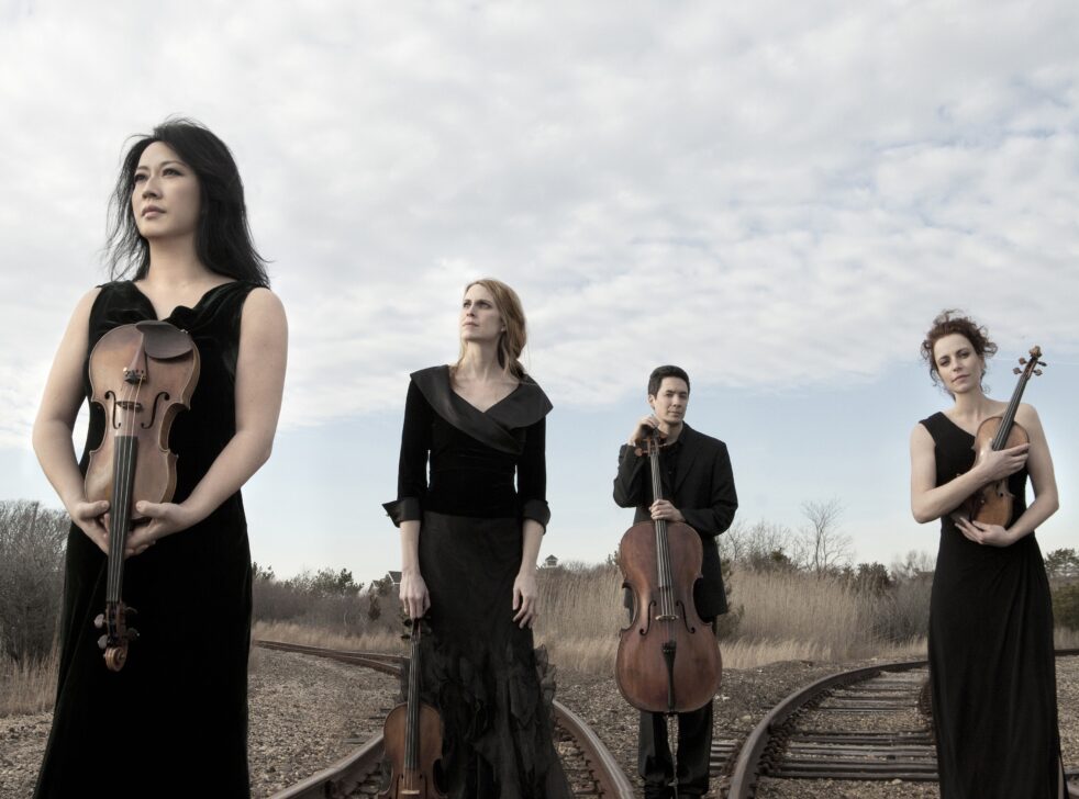 Four members of the Daedalus Quartet stand with their instruments on train tracks.