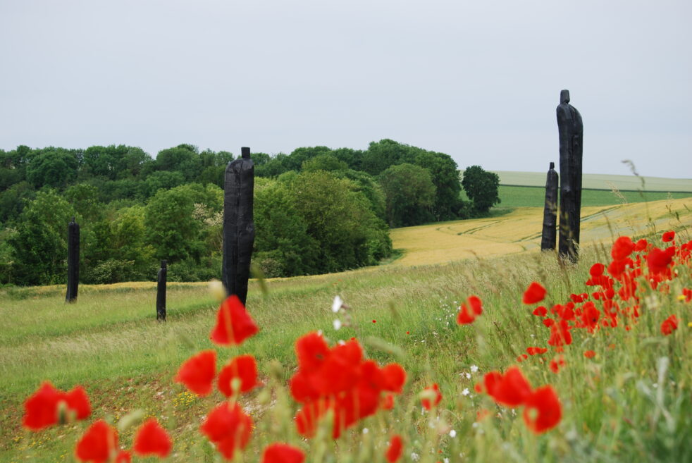 Tall, skinny, dark sculptures rising up out of a field on a hill, with red flowers in the foreground.