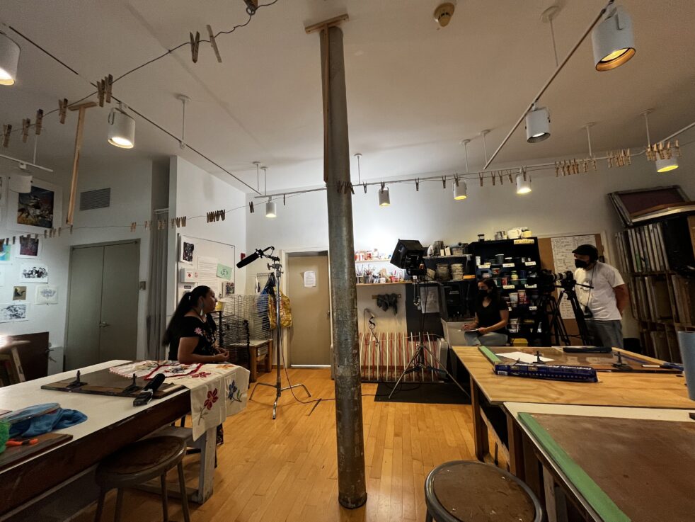 Three people in an interior space, two situated behind camera equipment and one sitting in front of the camera.