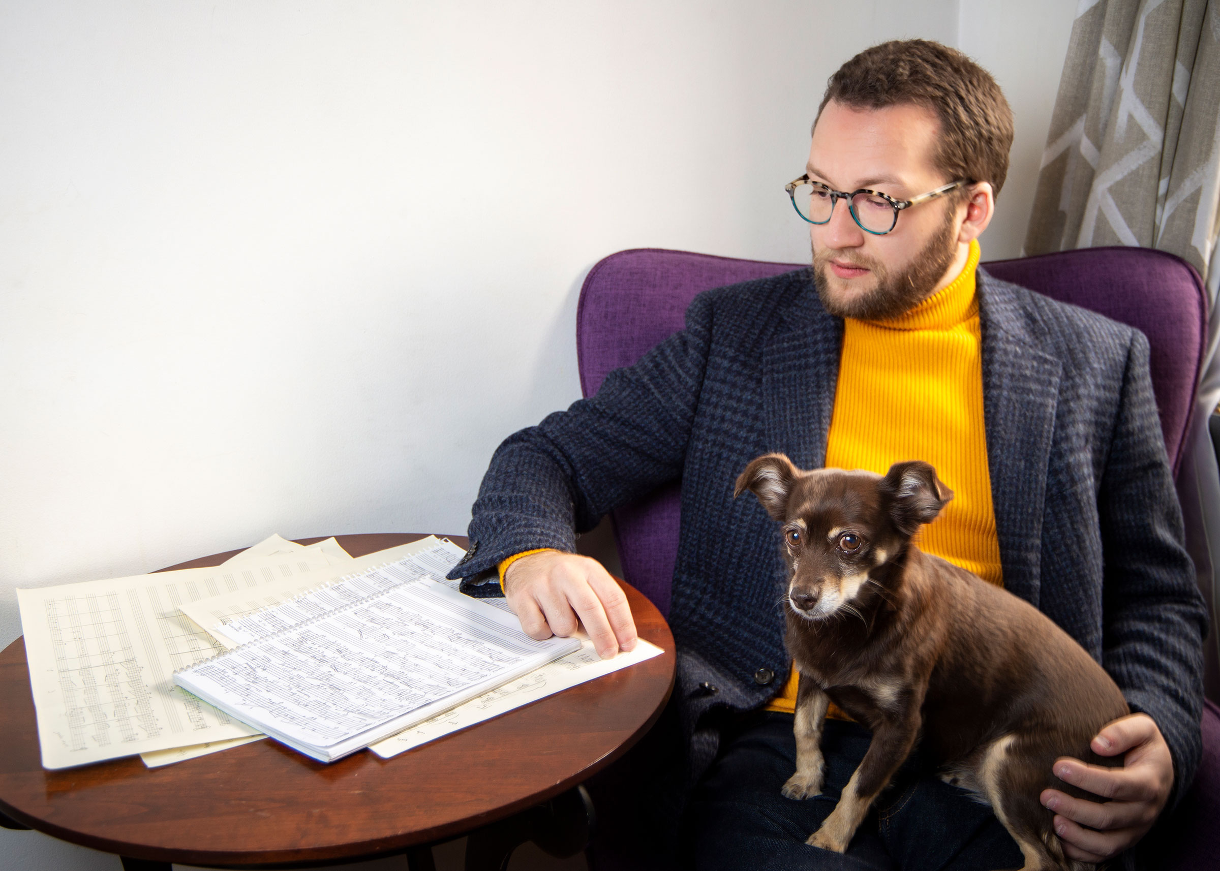 Man in glasses and yellow turtleneck with a small dog on his lap, looking at sheet music on a table.