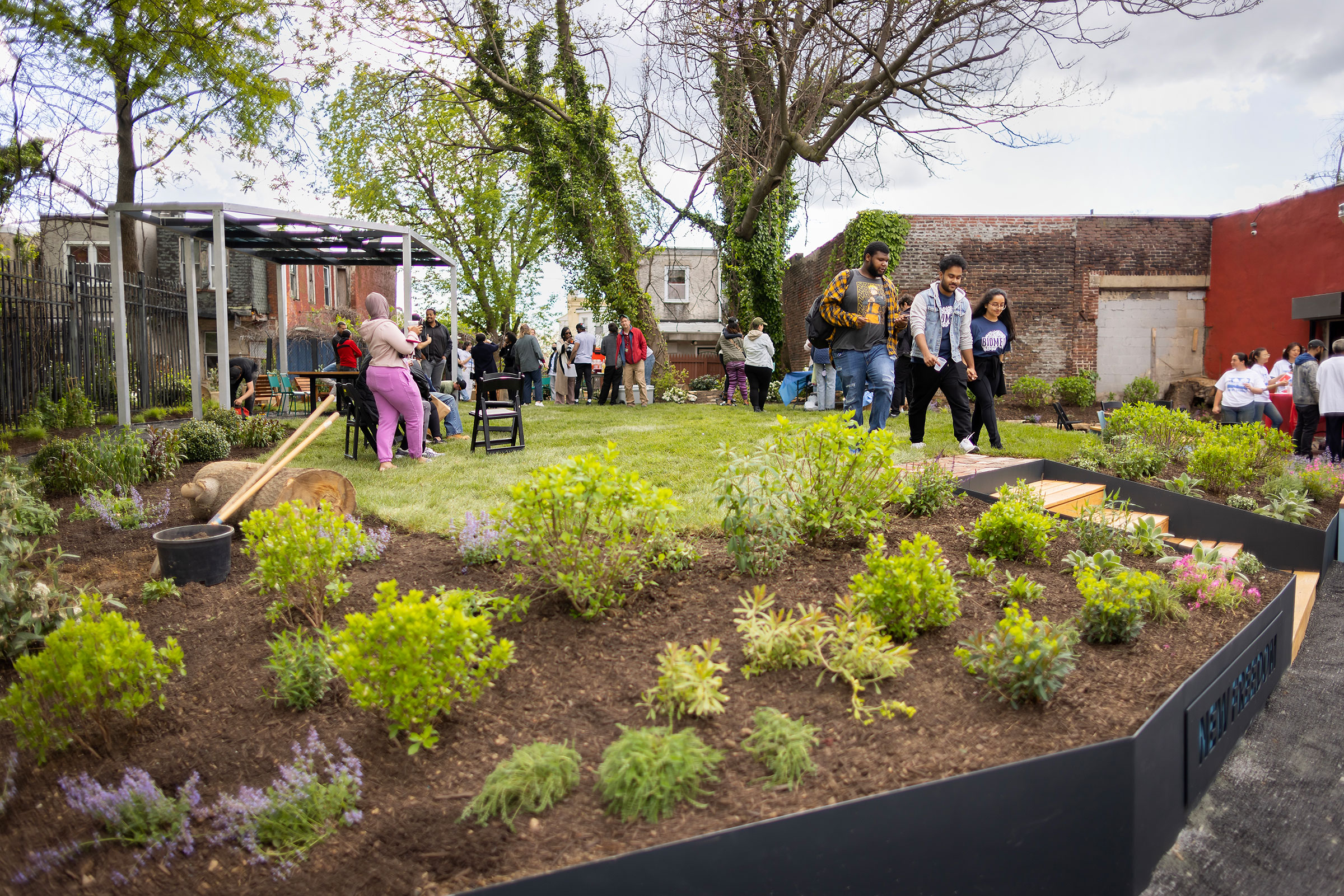 A community garden with people socializing, a pergola, and garden beds with green plants and purple flowers.