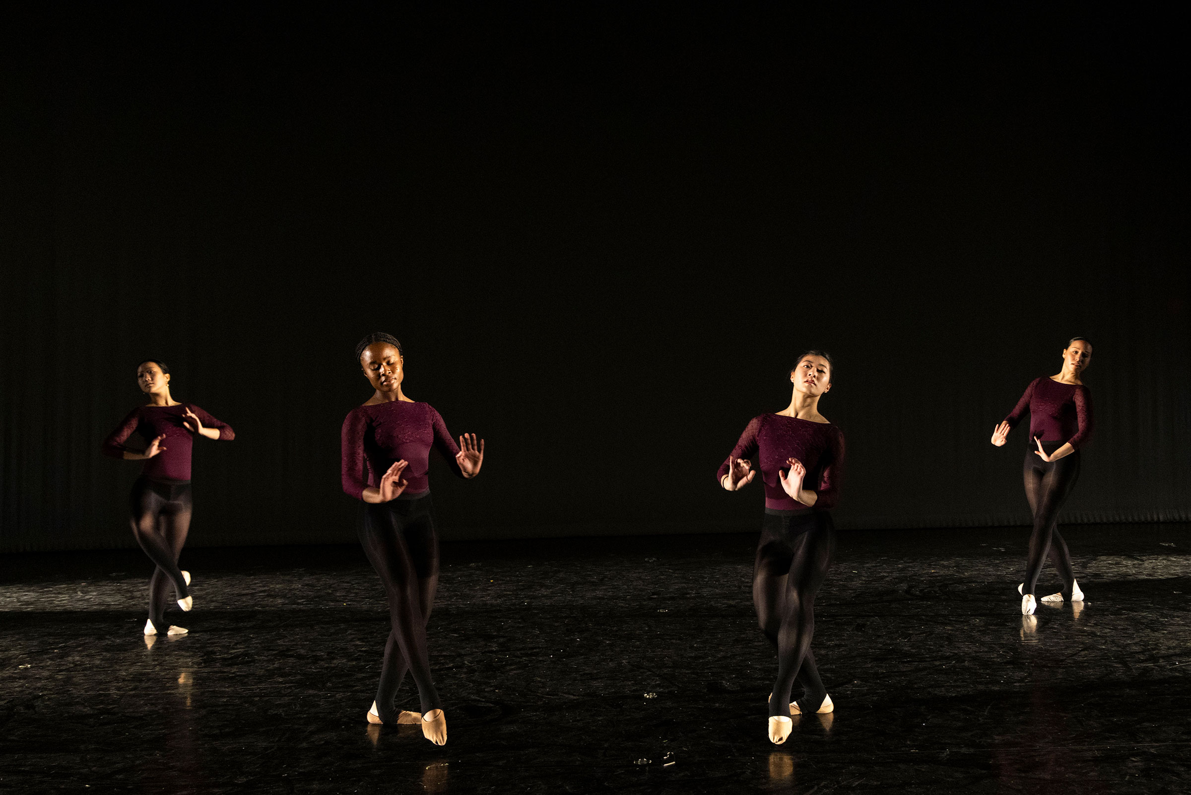 Four dancers on a dimly lit stage in maroon tops and black leggings, performing with raised arms.