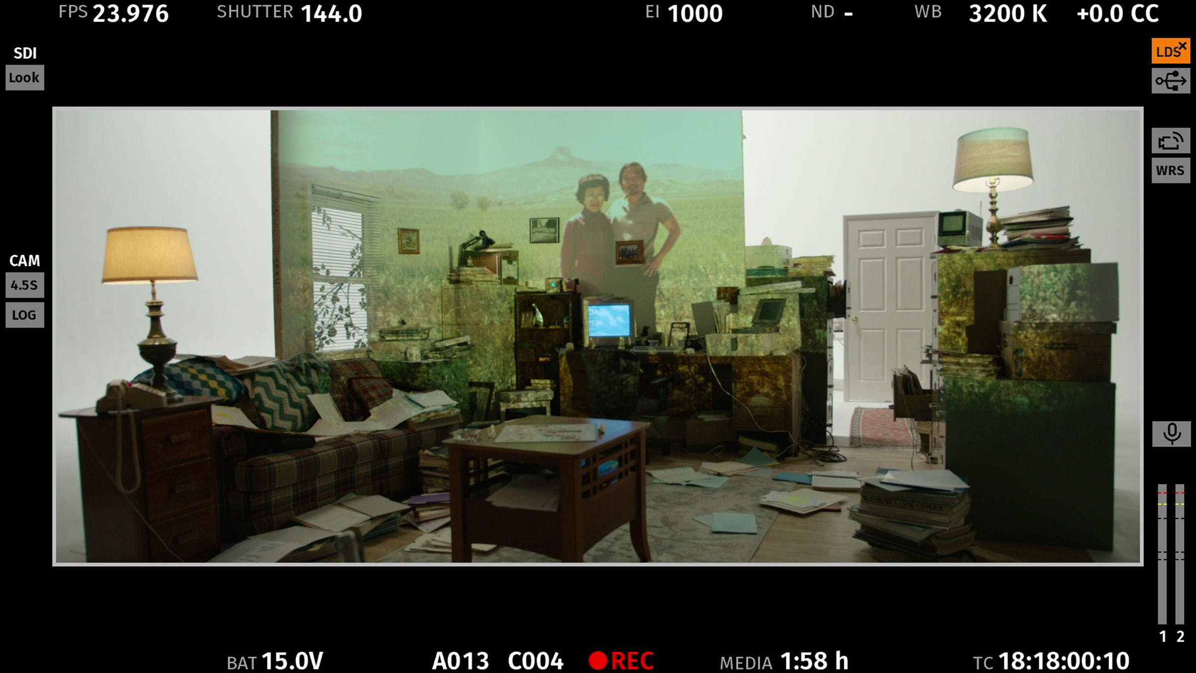 Domestic living room with a plaid sofa, scattered papers, and a projection of two people in a mountain landscape on the wall.