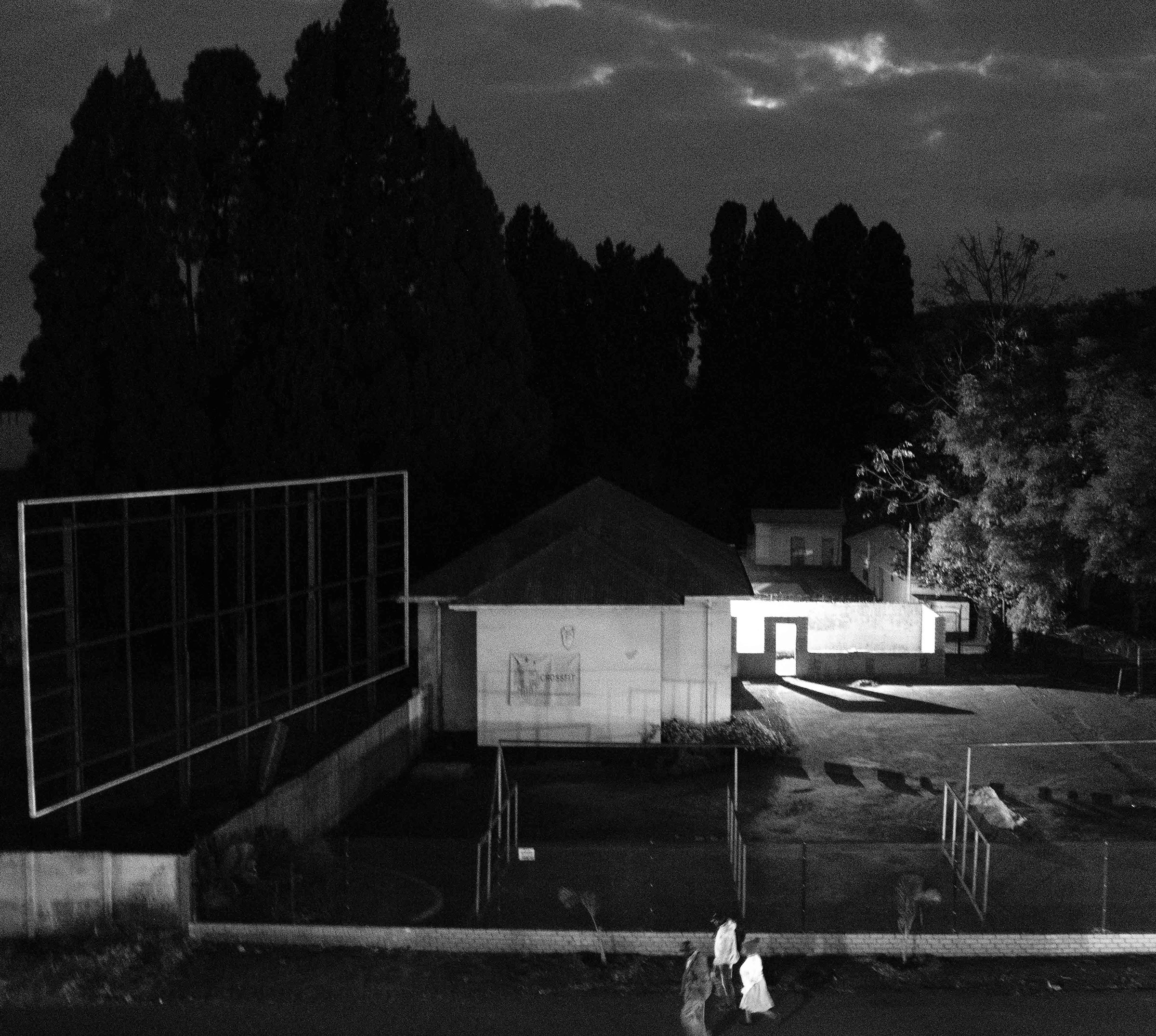 SithaboMathe_ViewfromThando'swindow_2400x2151.jpg Image Breakdown Long Description: The black-and-white image depicts an outdoor scene at night. In the foreground, there are two people walking along a paved area, illuminated partially by streetlights. To the left, a large, empty billboard frame stands against a backdrop of tall, dark trees. Central to the image is a simple, rectangular building with a slanted roof. Its walls are white and reflect soft light, with a small amount of text visible. Additional structures are seen to the right, partially obscured by shadows and trees. The sky is cloudy, with faint light peeking through. Alt-text: Nighttime scene with a building, billboard frame, and two people walking.