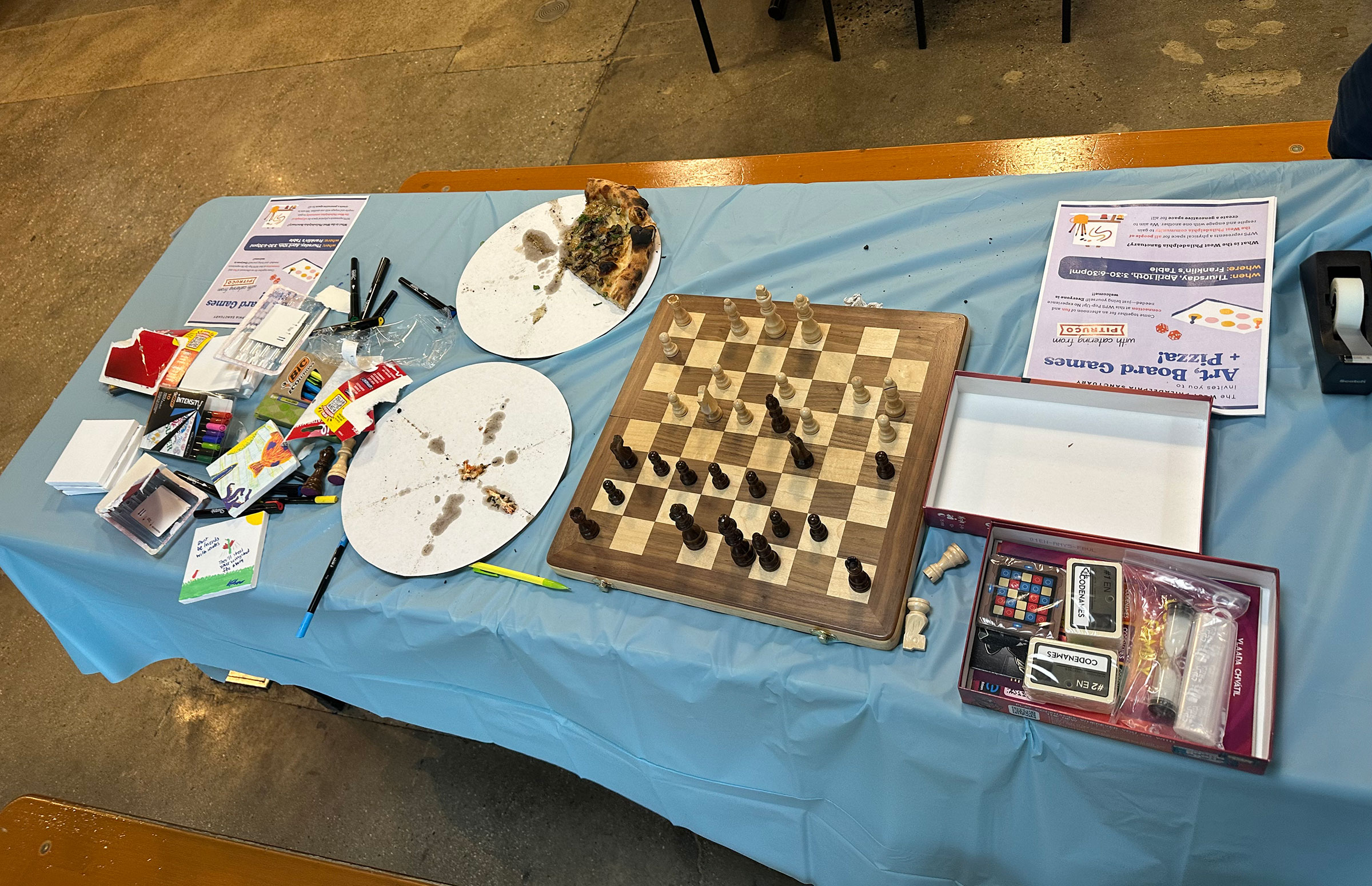 A table with art supplies, pizza trays, a chessboard, and board game components.