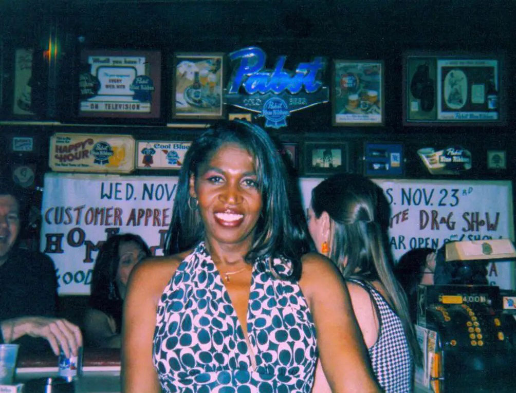 A smiling woman in a patterned dress standing in a bar with a neon "Pabst" sign in the background.