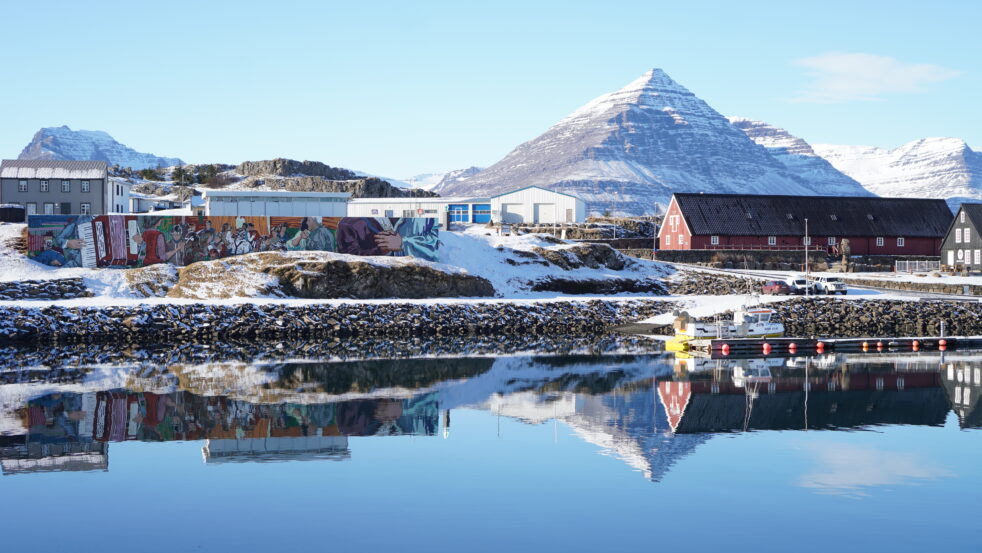 A waterfront icelandic town with colorful murals, snow-covered buildings, and mountains reflected in a calm body of water under a clear sky.