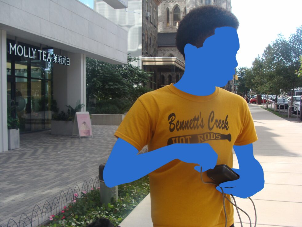 A person with blue arms and face in a yellow T-shirt with text stands near a building on a city street