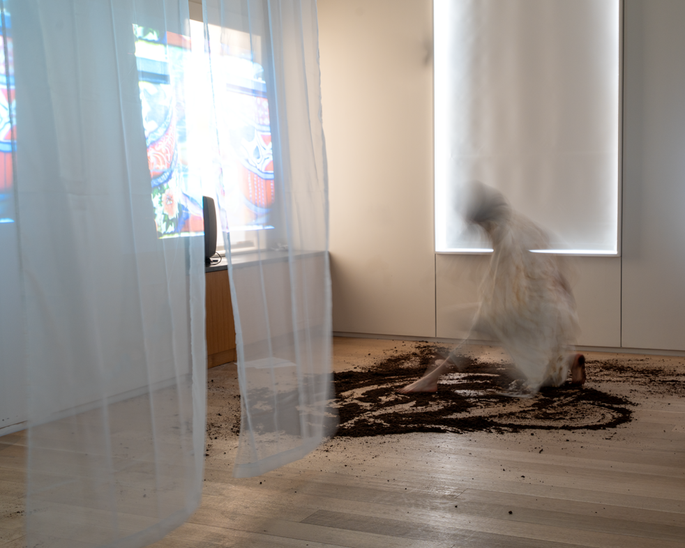 A blurred figure kneels on a wooden floor with dark soil spread around, partially obscured by a white curtain revealing a a projection resembling a stained glass window.