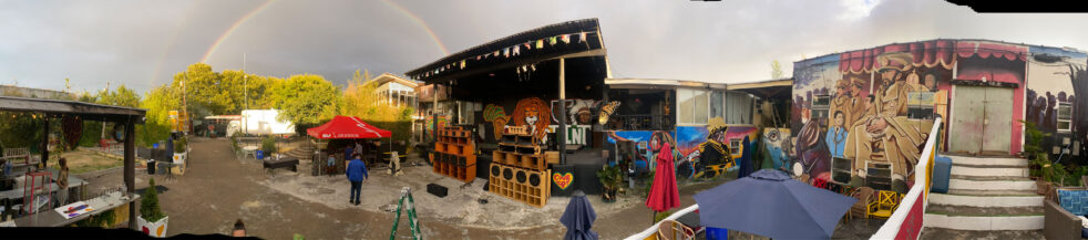 A panoramic view of a colorful outdoor music venue with sound system and rainbow visible.
