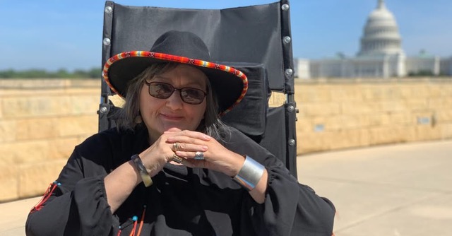 a woman in silver jewelry, sunglasses and beaded hat is seated outdoors in front of the US capital.