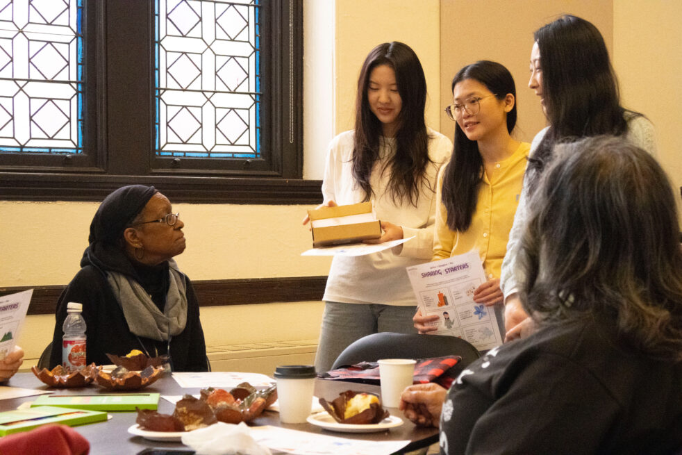People in a room, with some seated at a table and others standing, presenting with a box and a sheet titled "Sharing Starters" in front of stained-glass windows.