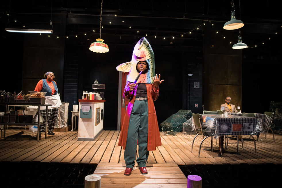 A theatrical stage with a person speaking towards the viewer in a fish costume while holding a knife, with other characters in a café setting background.